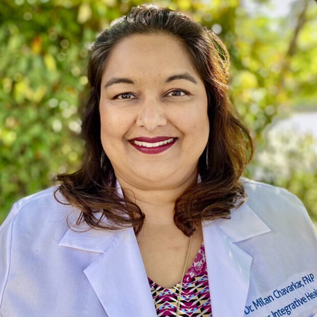 A woman wearing a white lab coat stands outdoors in front of green foliage, smiling at the camera. , Kiara Health Fertility