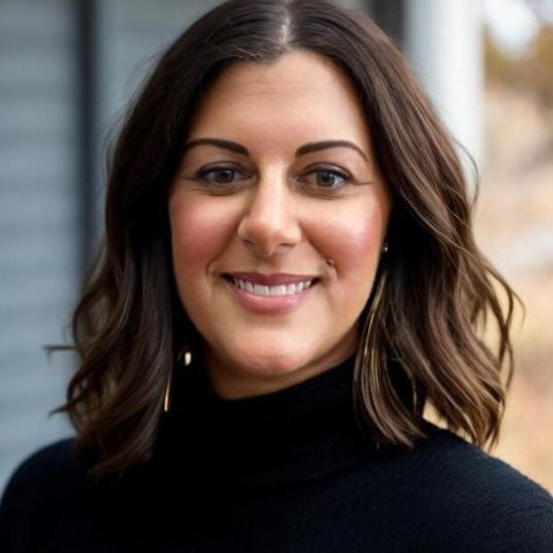 A woman with shoulder-length brown hair and a black turtleneck sweater smiles at the camera. The background is slightly blurred. , Kiara Health Fertility