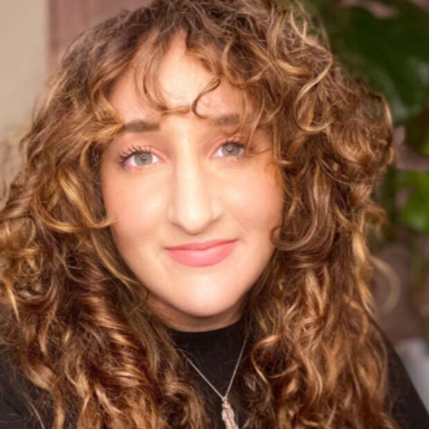 A person with long, curly brown hair and light eyes is smiling softly at the camera, wearing a black top. , Kiara Health Fertility