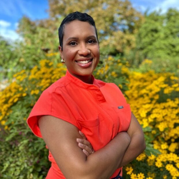 A woman wearing a bright orange top stands outdoors with arms crossed, smiling in front of a background of yellow flowers and green foliage. , Kiara Health Fertility