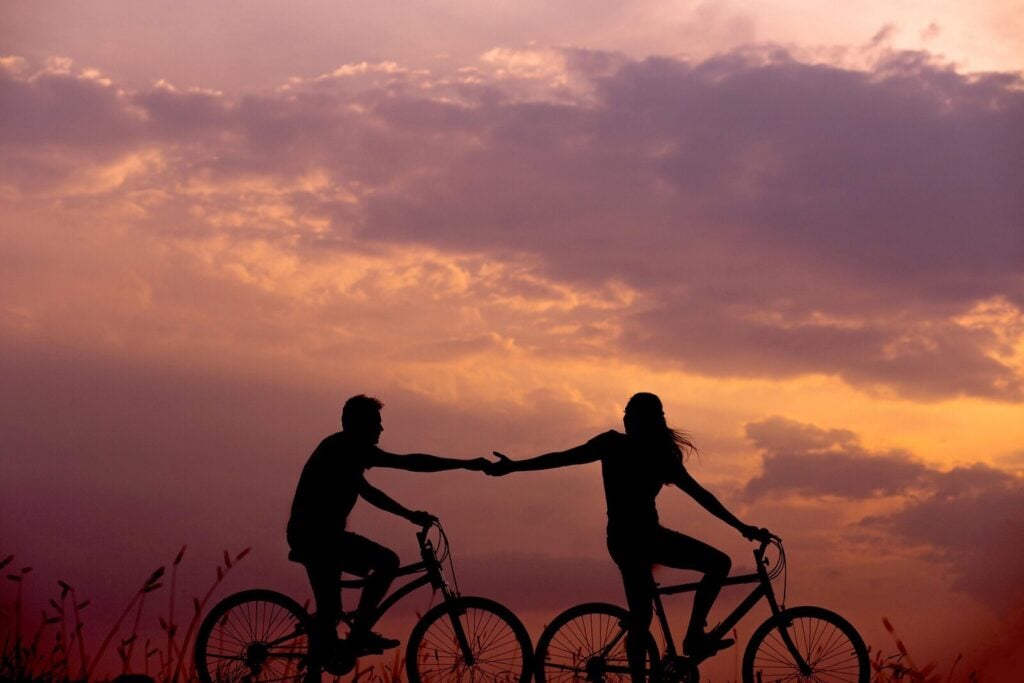 Two people riding bicycles at sunset, reaching out to hold hands with a cloudy sky in the background. , Kiara Health Fertility