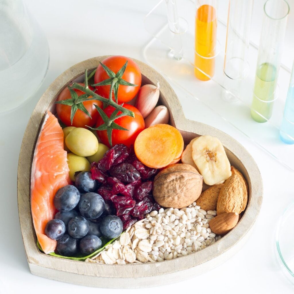 A heart-shaped wooden dish holds salmon, tomatoes, garlic, apricot, banana, nuts, oats, barley, blueberries, grapes, and dried cranberries—ingredients known to support nutrition and fertility—with test tubes in the background. , Kiara Health Fertility