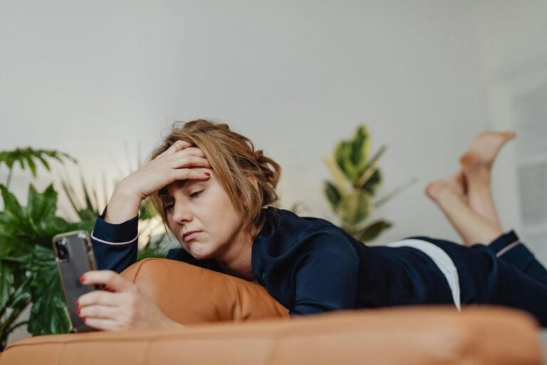 A woman in pajamas lies on a couch, holding her head with one hand and looking at her smartphone with a concerned expression. , Kiara Health Fertility