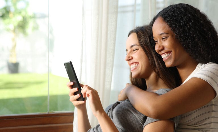 Two women sit closely together on a couch, smiling while looking at a smartphone. Sunlight streams through the window behind them, creating a warm atmosphere as they explore Kiara health tips together. , Kiara Health Fertility
