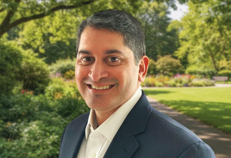 A man in a dark suit and white shirt stands outdoors in a park with green trees and a sunny background, smiling at the camera, reflecting the vibrant spirit of Kiara Health. , Kiara Health Fertility