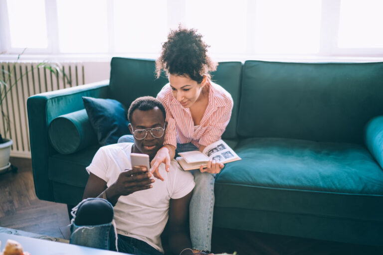 A man sits on the floor using a smartphone while a woman leans over him holding an open book; they are in a living room with a green sofa. , Kiara Health Fertility