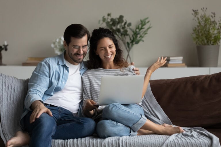 A man and woman sit closely together on a couch, smiling and looking at a laptop screen, appearing to enjoy a video call or online content. , Kiara Health Fertility