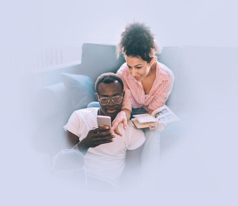 A man sitting on the floor looks at a smartphone while a woman beside him, holding an open book, looks on. They appear relaxed in a home setting, possibly exploring fertility support options together. , Kiara Health Fertility