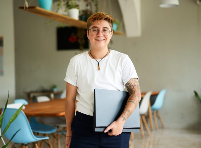 Person with short hair, glasses, and tattoos stands indoors holding a closed laptop, wearing a white t-shirt and dark pants. Tables, chairs, plants, and materials related to fertility support are visible in the background. , Kiara Health Fertility