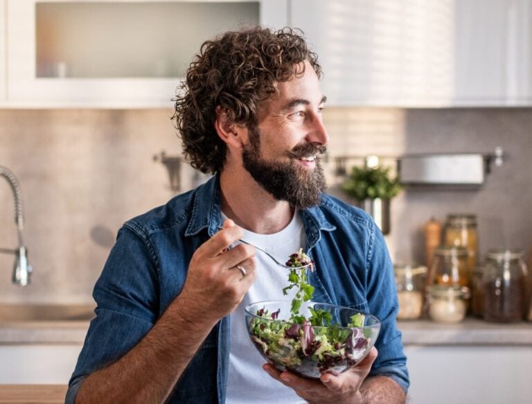 Man with curly hair and beard sits on kitchen counter, holding a fork and eating salad from a glass bowl, enjoying a moment of fertility support while looking out of a sunlit window. , Kiara Health Fertility