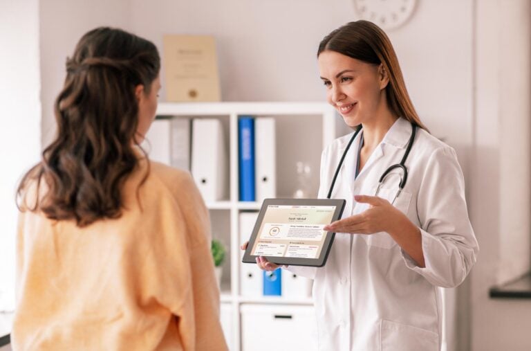 A doctor in a white coat from Kiara Health shows a patient information on a tablet in a medical office. The doctor is pointing at the screen and smiling during the consultation. , Kiara Health Fertility