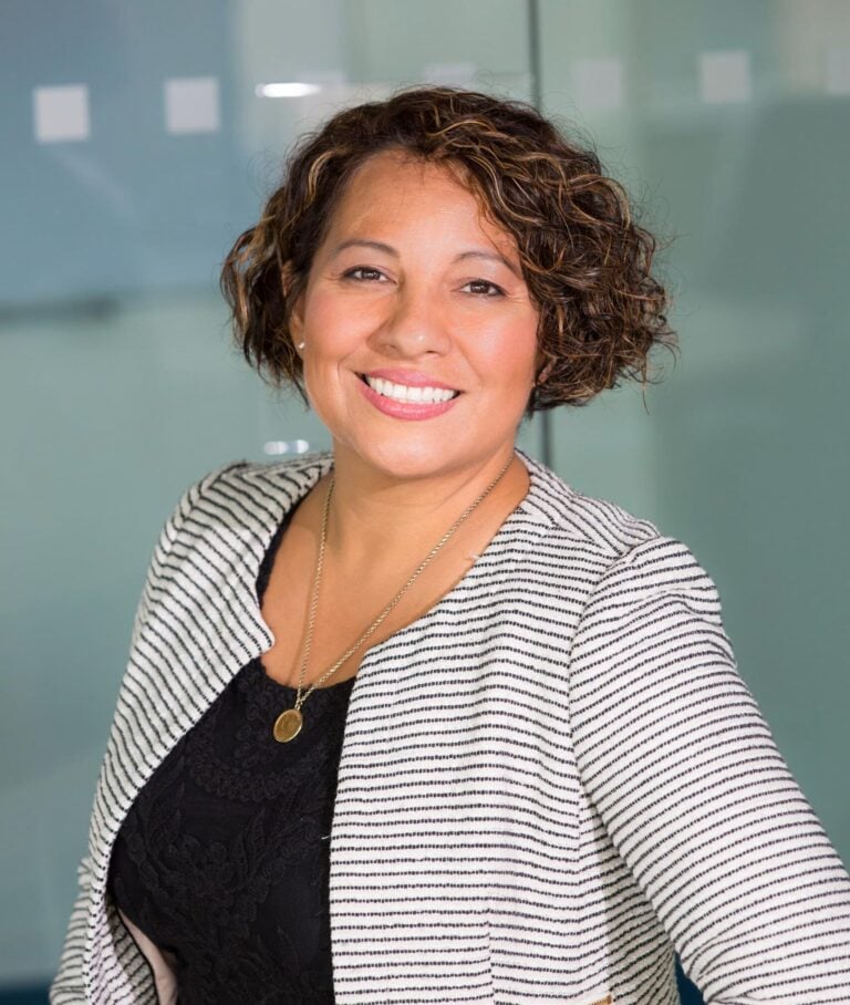Woman with short curly hair, wearing a striped blazer and black top, smiling while seated in a modern Kiara Health office setting with a glass wall in the background. , Kiara Health Fertility