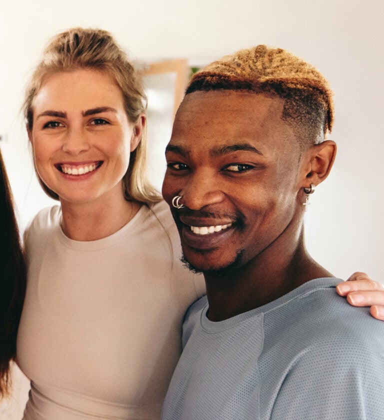 Two people stand close together indoors, smiling at the camera. One has light skin and long blonde hair; the other has medium skin and short blond-tipped hair with facial piercings. Their cheerful expressions radiate positivity, reflecting a vibrant sense of well-being and Kiara health. , Kiara Health Fertility
