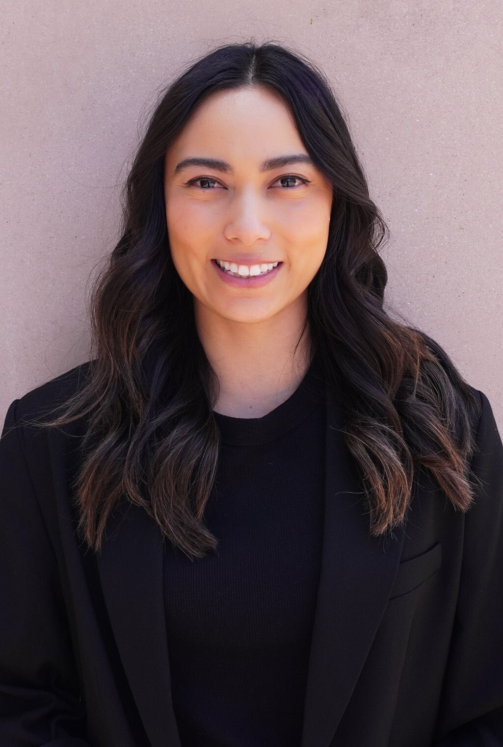 Woman with long dark hair wearing a black blazer and black shirt, smiling at the camera while standing against a light-colored wall—inviting you to discover more about her Free Coaching Session. , Kiara Health Fertility
