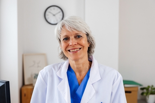 Smiling woman with short gray hair wears a white lab coat and blue scrubs, standing indoors with a clock and framed art in the background—an ideal image for your Landing Page, conveying professionalism and warmth. , Kiara Health Fertility