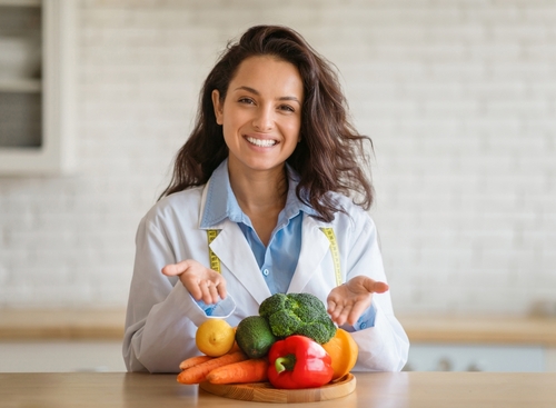 Woman in a lab coat sits at a table, smiling and gesturing toward assorted fresh vegetables and fruits on a wooden board—an inviting scene perfect for any Landing Page dedicated to healthy living and nutrition. , Kiara Health Fertility