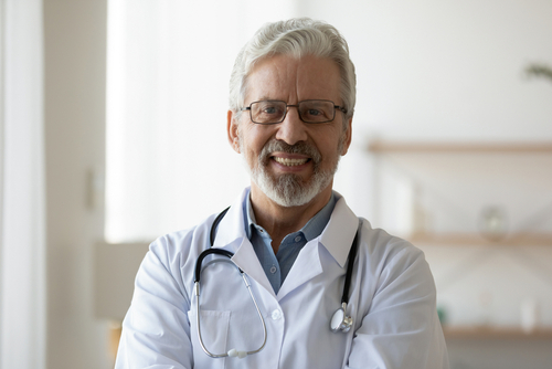 Smiling older male doctor with glasses, gray hair, and beard, wearing a white coat and stethoscope, standing indoors in a brightly lit room—ideal for featuring on a professional Landing Page or highlighting medical expertise for Landing Page 7.2. , Kiara Health Fertility