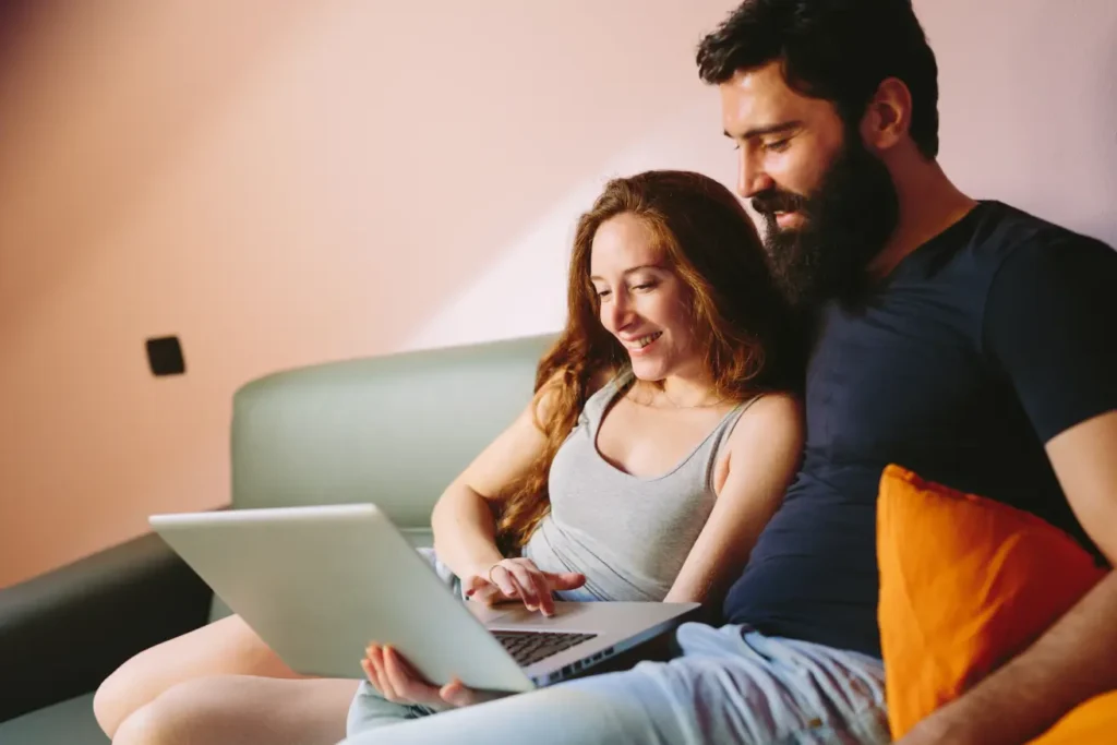 A woman and a man sit on a couch looking at a laptop together, exploring the features of Landing Page 7.2. The woman is smiling and using the keyboard, while the man has his arm behind her and looks at the screen. , Kiara Health Fertility