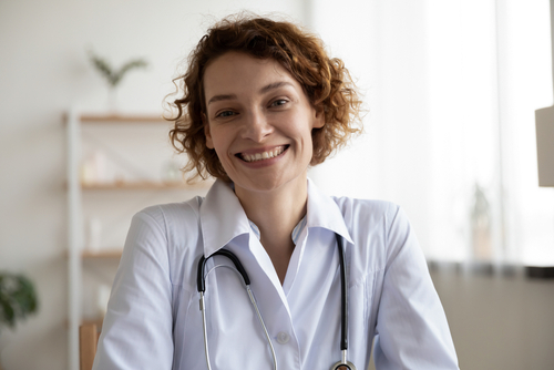 Smiling woman wearing a white lab coat and stethoscope sits indoors, looking at the camera—perfect for use on a professional Landing Page or as part of Landing Page 7.2 design concepts. , Kiara Health Fertility