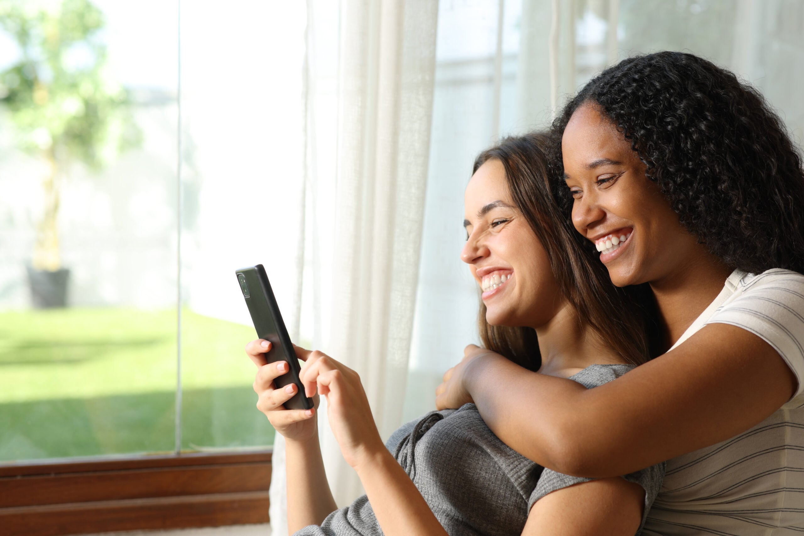 Two women sit by a window smiling and looking at a smartphone together, with one hugging the other from behind, their faces lit up by the excitement of exploring the latest Kiara Advanced features. , Kiara Health Fertility